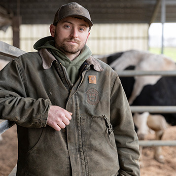 250107-Harmonyway-Farm-051.jpg A young male farmer poses for a photo in front of a cow.